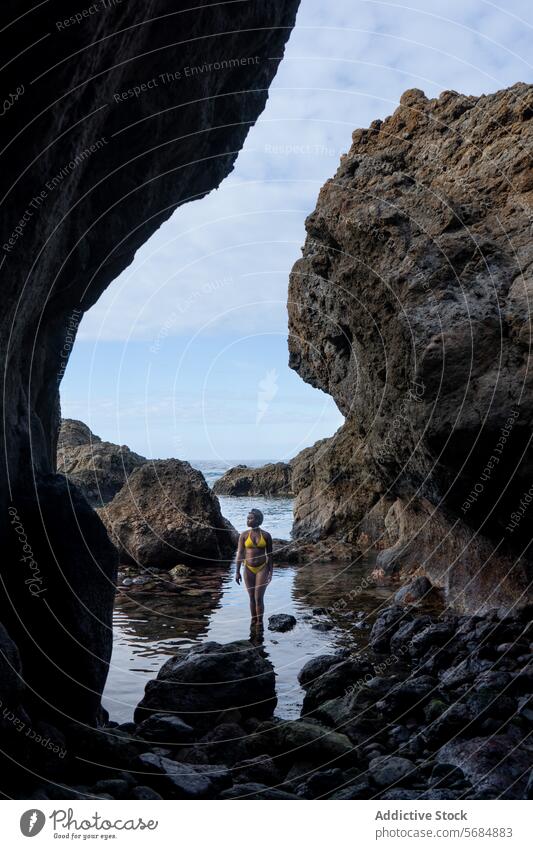 Young black woman in bikini standing on stone tourist cave rock swimwear rocky cliff volcanic formation female young sea nature coast blue sky ocean cloudy