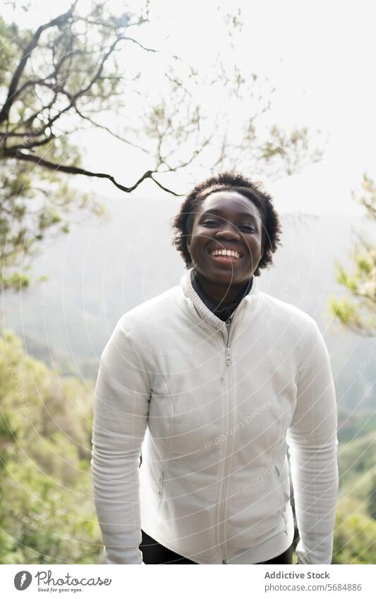 Cheerful black young woman standing against green trees traveler smile forest fog cheerful mist woodland nature female african american ethnic jacket happy park