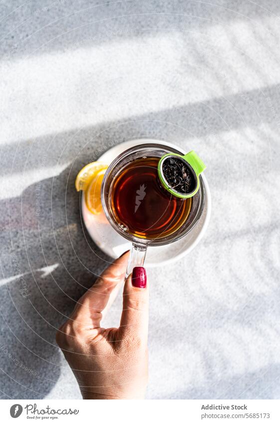 From above of anonymous hand holding a clear cup of tea with a green infuser and a lemon slice on the side, casting shadows on a textured surface overhead glass