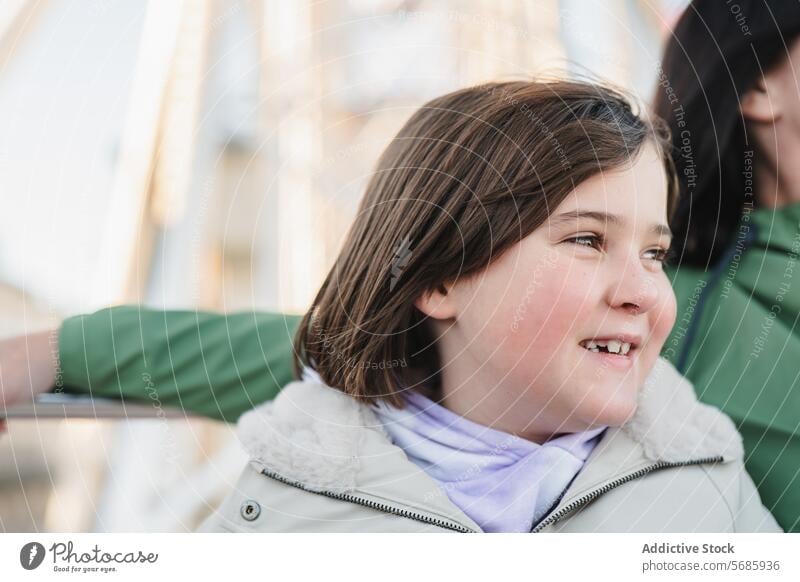 A girl in a beige coat with a dreamy expression looks up while sitting in front of a Ferris wheel amusement park child contemplative happy casual outdoor