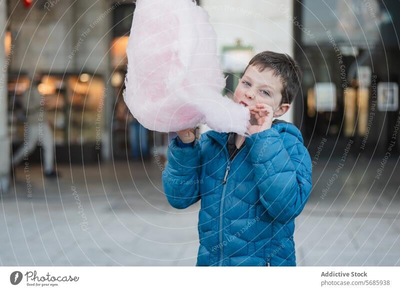 A boy in a blue jacket looking at camera while biting into a fluffy pink cotton candy taste snack confection sweet sugary treat dessert indulgence enjoyment