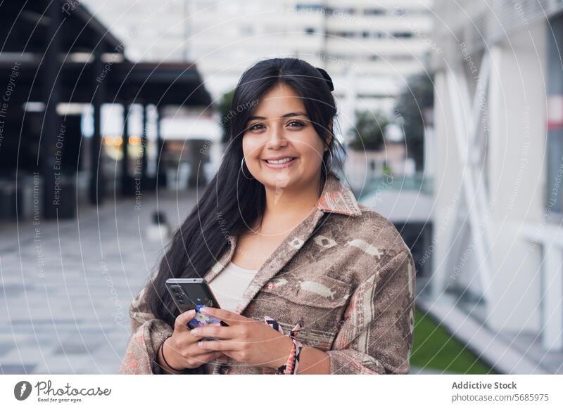 Happy woman using smartphone near glass wall at city confident jeans sneakers smile happy street modern style urban female gadget building device lifestyle lady