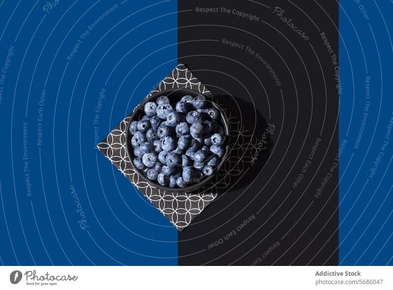 Top view of bowl of juicy blueberries on a geometric patterned napkin, set against a contrasting blue and black diagonal background Blueberry fruit ripe food