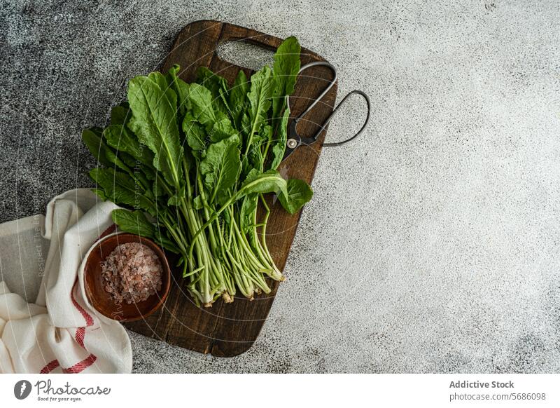 Top view of bunch of fresh arugula herb leaves on a rustic wooden cutting board, accompanied by a bowl of pink salt and kitchen shears, set on a grey textured surface