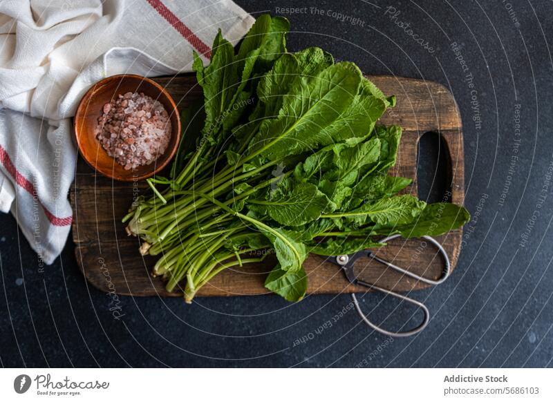 Top view of bunch of fresh arugula leaves alongside a bowl of pink salt and scissors on a rustic wooden cutting board, set against a dark tabletop with a striped towel