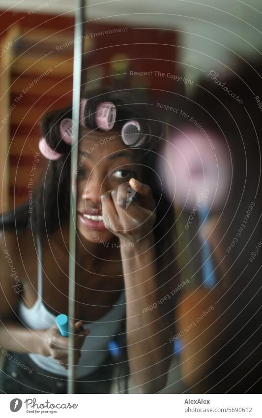 Young, long-haired, slim woman with curlers in her hair applies make-up to her face in front of a split mirror Woman Young woman Long-haired Dark Feminine