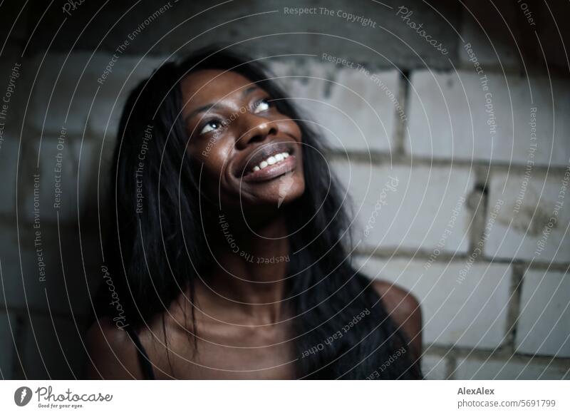 Young, long-haired, slender woman stands in front of a wall of white bricks, looks up at the light and smiles Woman Young woman Long-haired Dark Feminine