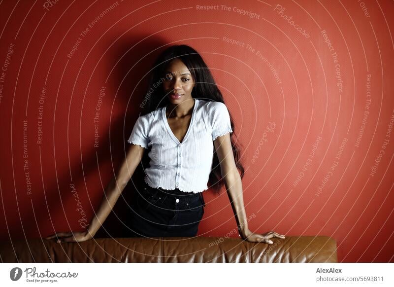 Young, long-haired, slim woman stands leaning on a sofa in front of a red wall and looks past the camera Woman Young woman Long-haired Dark Black Feminine