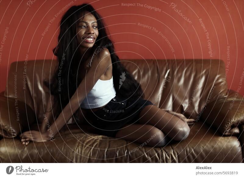 Young, slim woman with long black hair and hot pants sits crosswise on a brown couch in front of a red wall and smiles into the camera Woman Young woman