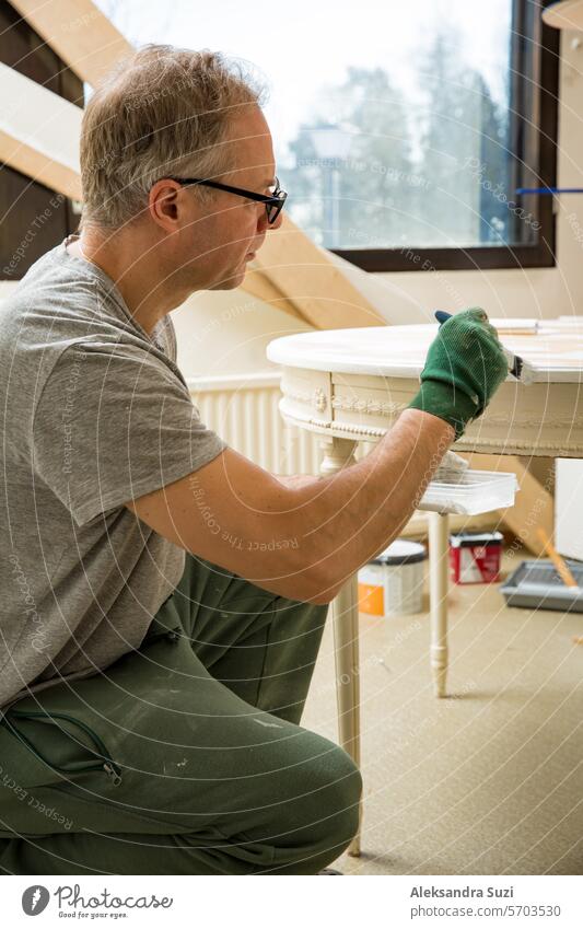 Middle-age man painting old wooden table with paint brush in white color. Furniture restoration, DIY repair, repair by oneself and home improvement. Recycling and sustainable lifestyle