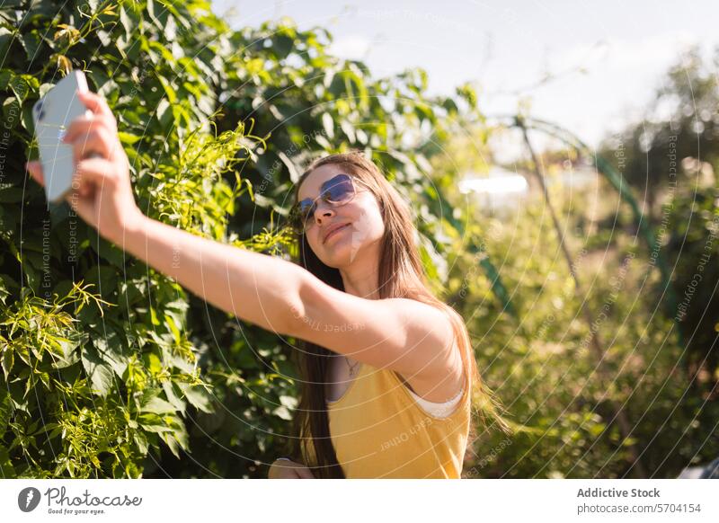 Woman taking selfie in sunny green garden woman phone greenery yellow tank top cheerful young lush day smartphone outdoor lifestyle nature casual leisure