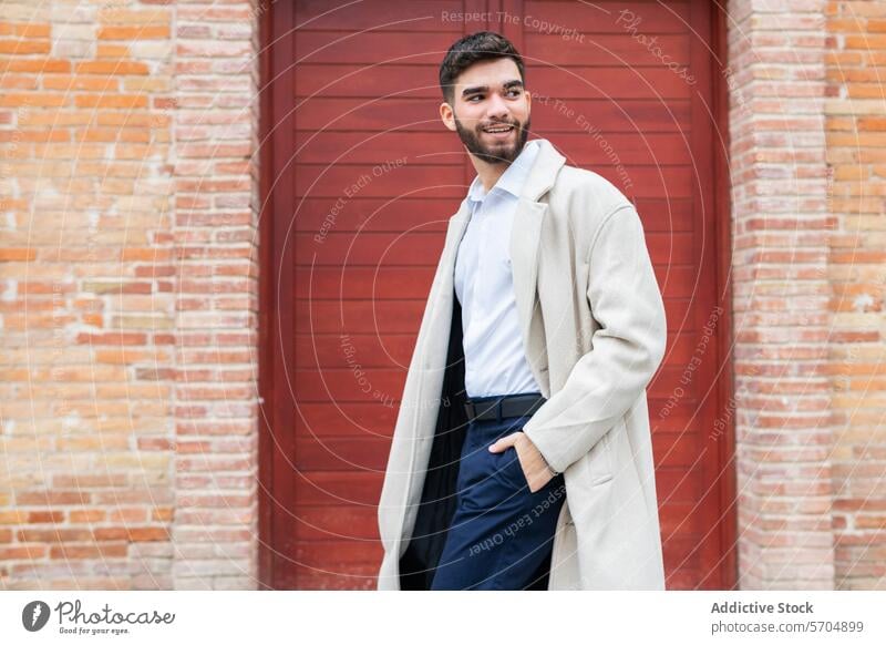 A stylish young man in business attire smiles confidently while standing against a red door backdrop, exuding a sense of casual professionalism in his beige coat and navy trousers