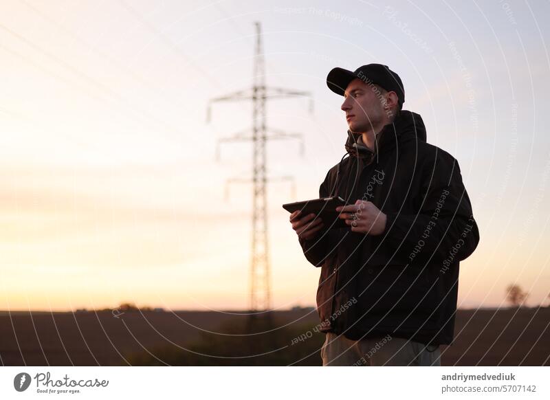 Young power engineer holds digital tablet against high voltage tower at field. Electrical engineer checking energy efficiency electrical towers. World Energy Saving Day. Energy business concept.