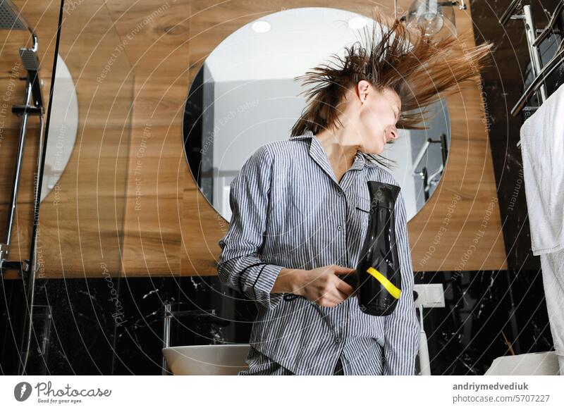 Concept of haircare and having fun in bathroom. Bottom view of cheerful beautiful woman singing while drying hair with blowdryer and using a hair dryer as a microphone at home stylish bath interior