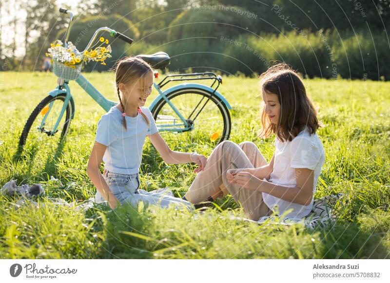 Two teenage girls spend time on green grass lawn in park, braid pigtails and tails for each other, enjoy summer and vacations attractive beauty bicycle bike