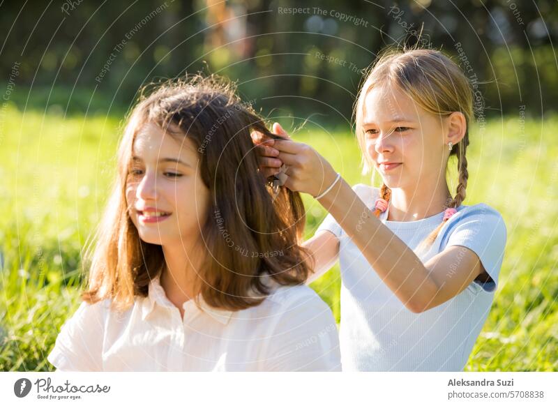 Two teenage girls spend time on green grass lawn in park, braid pigtails and tails for each other, enjoy summer and vacations attractive beauty bicycle bike