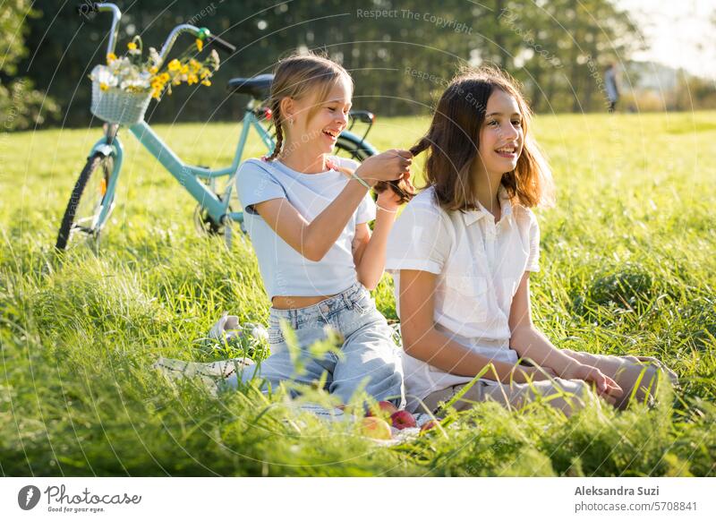 Two teenage girls spend time on green grass lawn in park, braid pigtails and tails for each other, enjoy summer and vacations attractive beauty bicycle bike