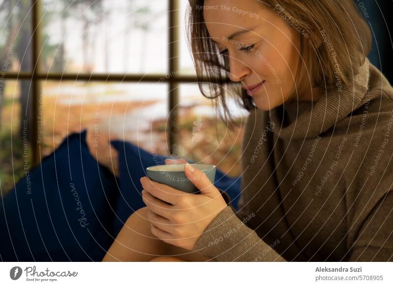 Pretty young woman reading book and drinking morning coffee at home sitting at the window behind which there is an autumn landscape adult background beautiful