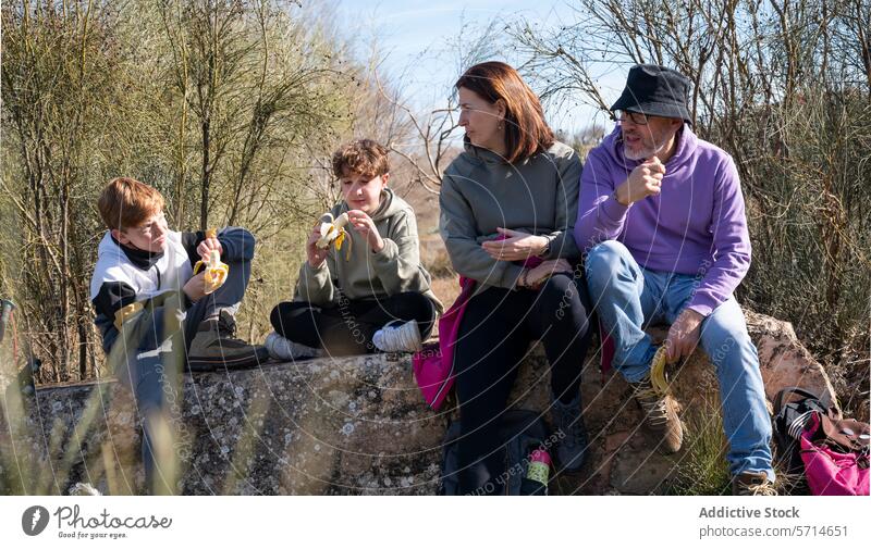 A family enjoys a snack break on a rustic stone bench during a hike, with two children peeling bananas and adults looking on leisure trail outdoor eating