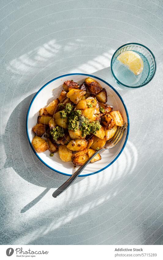 Top view of sunlit roasted potatoes with a dollop of green pesto on a white plate with blue trim, next to a glass of water with lemon, casting a shadow on a textured grey surface