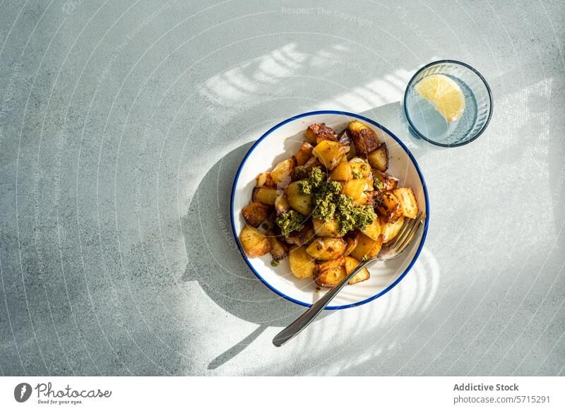 Top view of sunlit roasted potatoes with a dollop of green pesto on a white plate with blue trim, next to a glass of water with lemon, casting a shadow on a textured grey surface