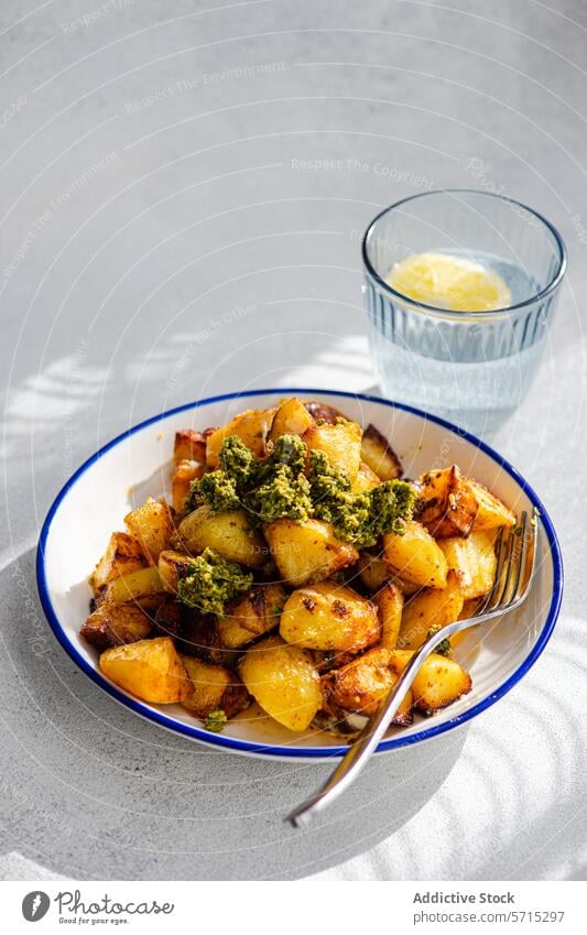 Roasted golden potatoes topped with vibrant green pesto on a white enamel plate with a blue rim, accompanied by a clear glass of water, all set upon a light grey textured surface