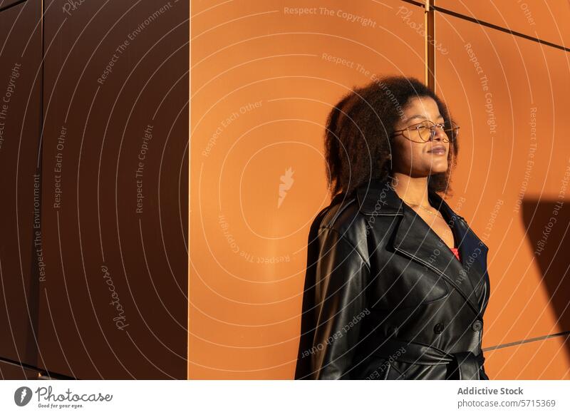 Contemplative woman in a black leather jacket stands before an orange wall, bathed in soft sunlight Woman black jacket contemplative winter lifestyle urban