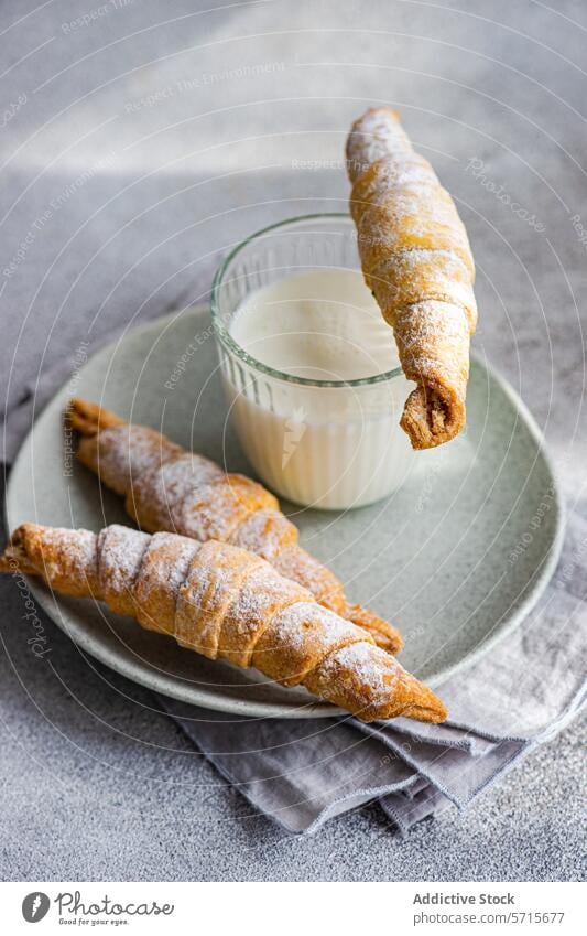 Top view of freshly baked homemade pastry cone filled with jam, partially dipped in a glass of milk, with another pastry on a plate, on a grey textured surface