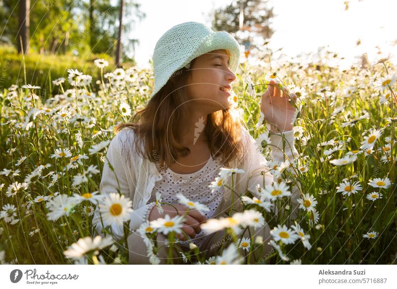 Girl sitting on a meadow covered with wild flowers. Background of field of daisy flowers in bloom. Sunny summer day, rays of light. Summer vacation concept