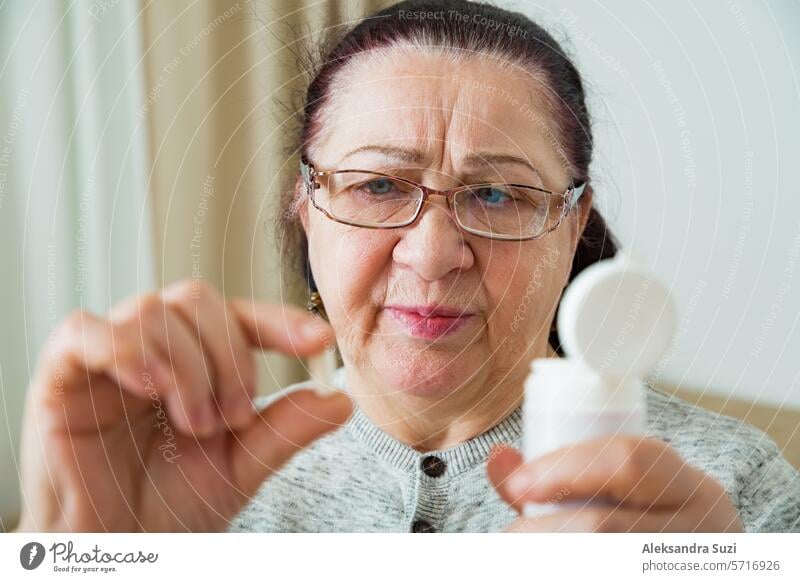 Senior Woman Taking Medication, holding pill in hand, looking though glasses. Elderly Person with concerned face reading prescription on medicine bottle. adult