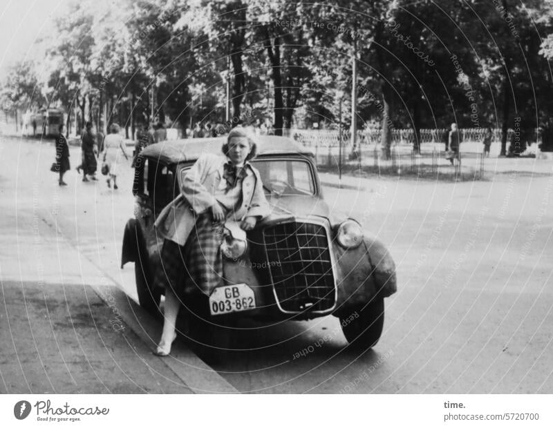 radiator mascot Woman portrait Looking into the camera Blonde car Park Street Ajar motor vehicle Road traffic Sidewalk trees Vintage car pose Dream cart Coat