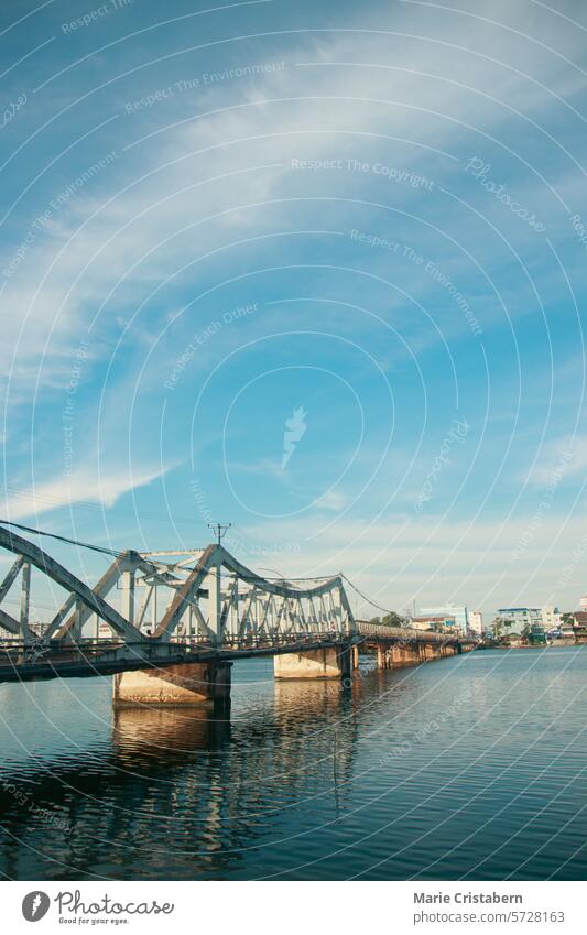 A calm river flows under a the famous old bridge in Kampot town in Cambodia, reflecting the clear blue sky and scattered clouds, with a cityscape in the background