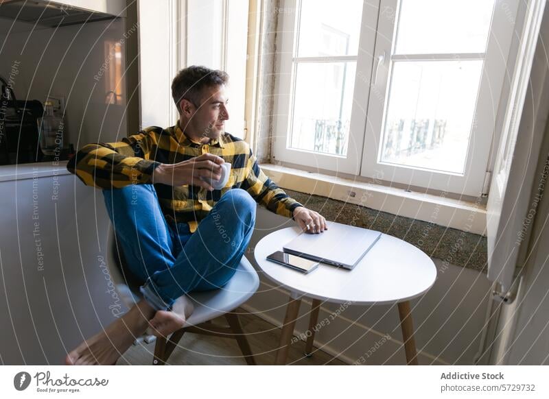 Contemplative man with coffee looking out the window contemplation break table sitting checkered shirt holding mug closed laptop smartphone indoor daylight