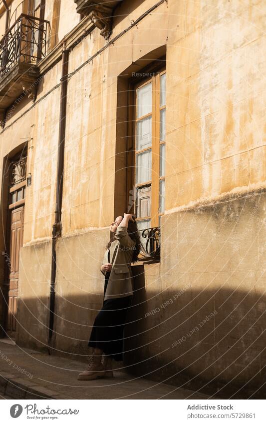 A young woman stands lost in thought, her hand gently touching her hair, in the quiet warmth of a sunlit historical alley contemplative urban tranquil building