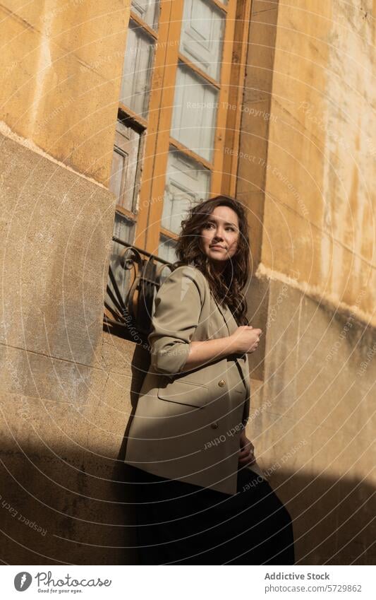 Captured in a beam of sunlight, a contemplative woman leans by a window on a textured old wall, exuding casual elegance reflective elegant fashion style coat