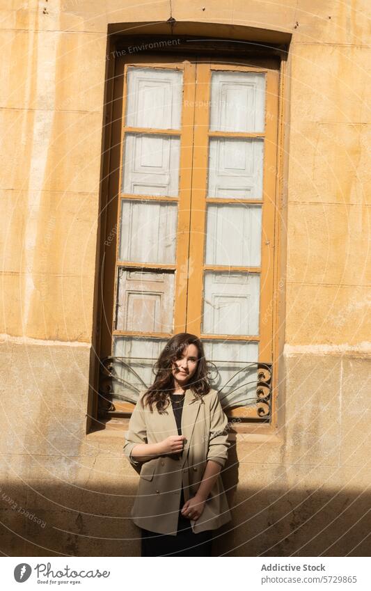 A fashionably dressed woman stands confidently in front of a vintage window, her expression serene in the warm sunlight chic style sunlit coat casual elegance