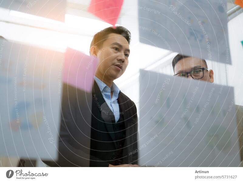 Two men are looking at a wall covered in paper with graphs and charts. They are discussing the information on the wall business businessman person office