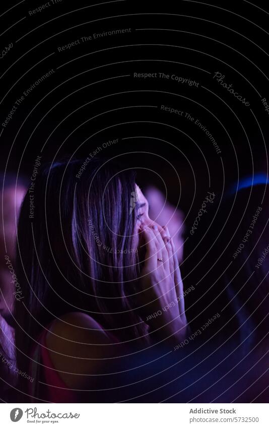 A woman in a crowd at a concert is captured in a profound moment of emotion, her hands covering her face against a backdrop of ambient stage lighting live