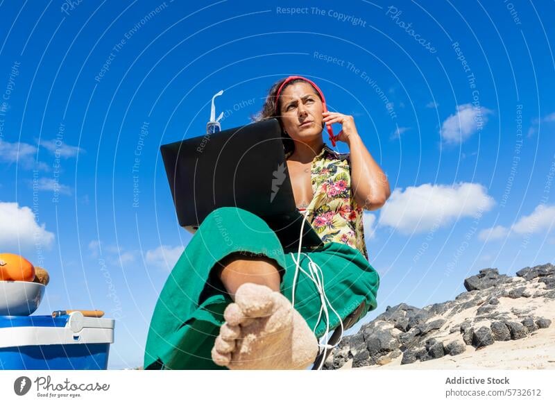 From below of focused digital nomad conducts a business call with a laptop on her knees, set against a backdrop of rugged beach rocks and blue sky woman