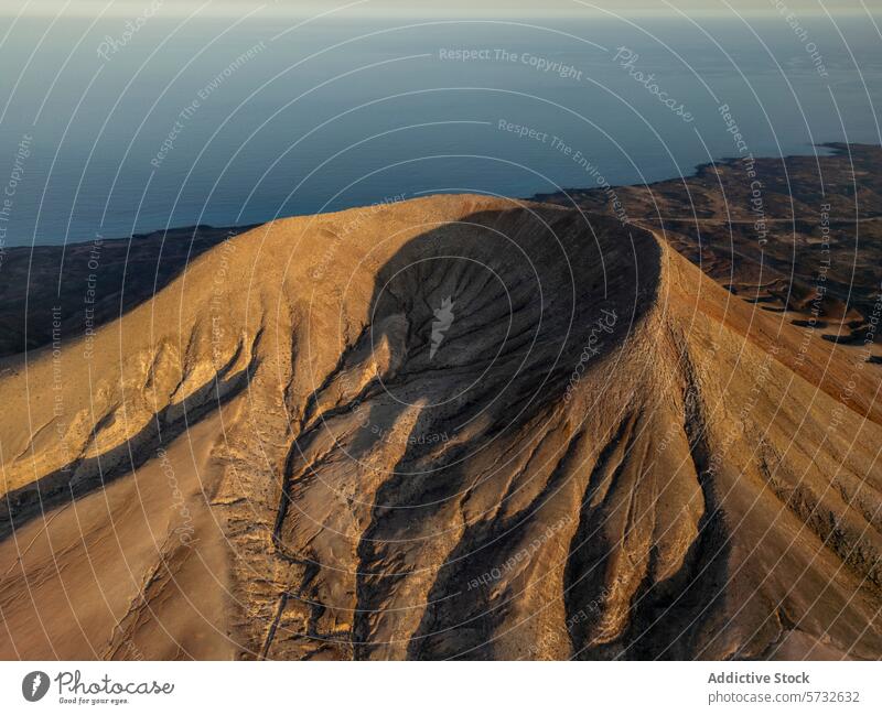 An aerial perspective captures the last rays of the sun casting shadows and golden highlights over a volcanic crater in Fuerteventura, with the ocean at a distance