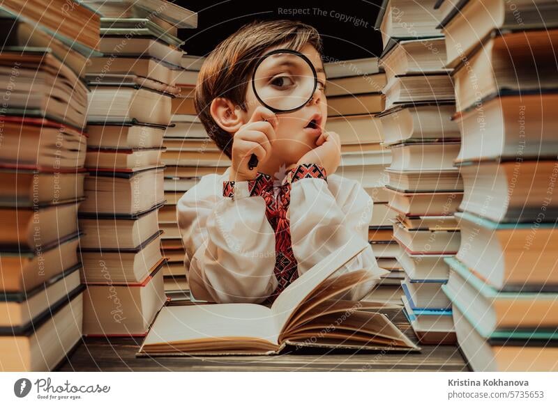 Little ukrainian researcher boy reads book with magnifying glass in library. Cute clever preschooler playing, studying knowledge with instrument education