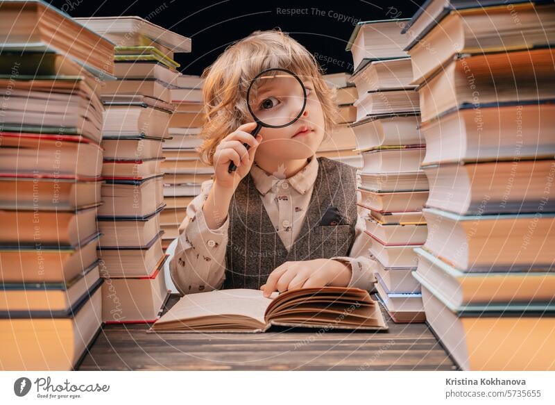 Little researcher boy reads book with magnifying glass in library. Cute clever preschooler playing, studying knowledge with instrument education literature