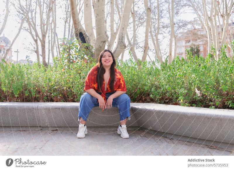 Smiling woman enjoying a sunny day in the park bench sitting smiling looking at camera cheerful city greenery casual clothing jeans fashion urban outdoor