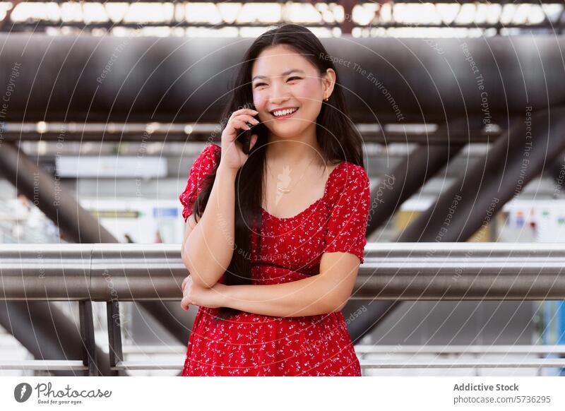 Smiling young woman talking on phone in urban setting mobile phone conversation smiling red dress railing metal blurred background city infrastructure leaning