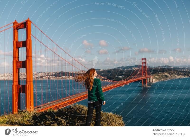 Back view woman stands in awe as the wind catches her hair, overlooking the magnificent Golden Gate Bridge on a vibrant spring day San Francisco breeze landmark