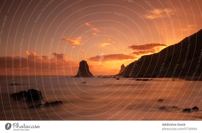 A fiery sunset sky casts a warm glow over the mystical sea stacks and tranquil waters at Playa de Benijo, Tenerife beach coastal seascape nature outdoor dusk