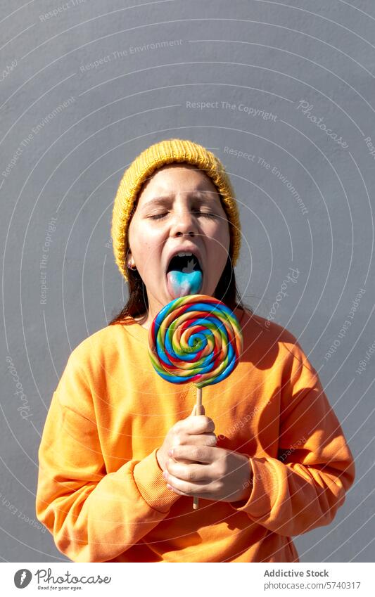 Young person enjoying a colorful lollipop young individual orange sweatshirt yellow beanie taste large swirl plain background enjoyment savoring vibrant candy