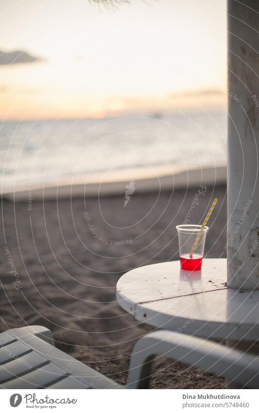 red cocktail in plastic glass at beach table by the ocean at dusk after sunset. Relaxing vacation. Summer drink. beverage Sandy beach Beverage Cocktail