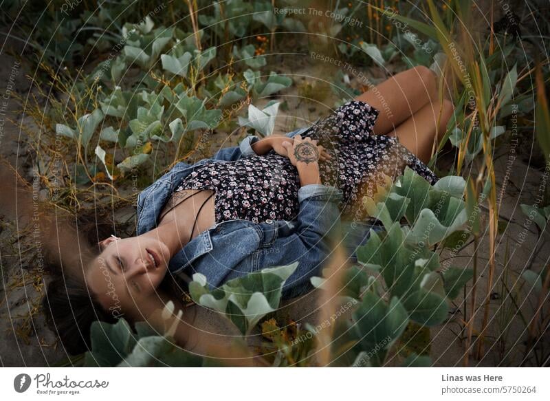 A stunning brunette woman in a blue denim jacket and summer dress is reclining on the sand amidst these enchanting dunes. Her skin is beautifully tanned, and adorned with some tattoos. She appears relaxed, lost in her thoughts.