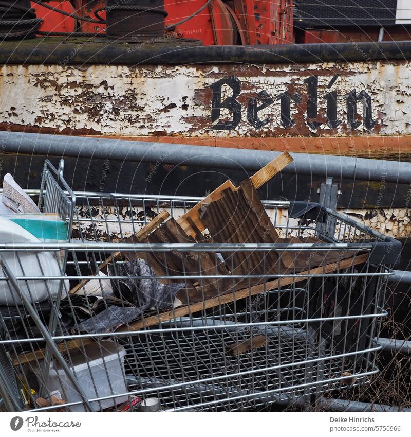 Shopping cart with garbage stranded in front of old barge labeled Berlin berlinerwasser City urban ship rusty Mobility turned off Shopping Trolley